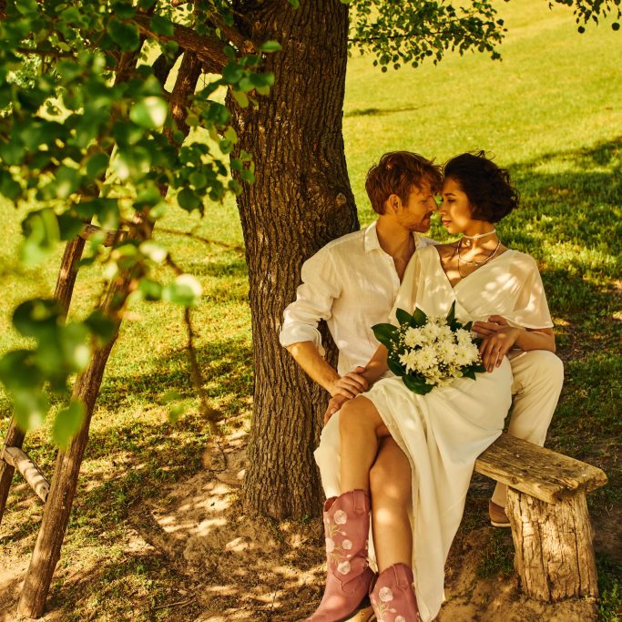 redhead groom embracing asian bride with bouquet while sitting on bench under tree, rustic wedding