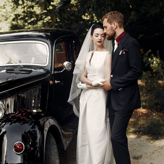 luxury elegant wedding couple gently embracing and holding hands on background of stylish black car. gorgeous bride and handsome groom in retro style. romantic moment